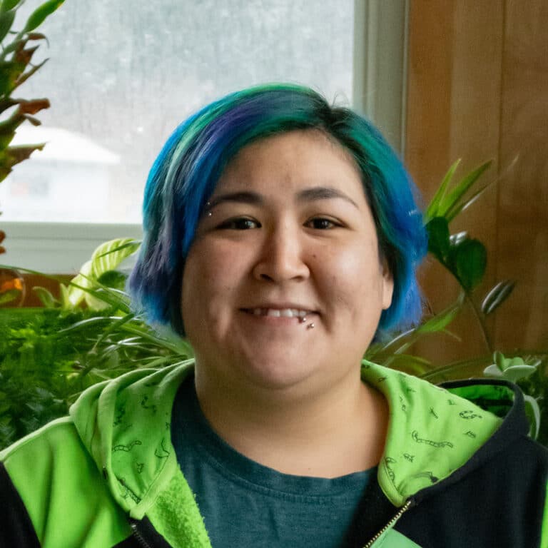 Woman with blue and green hair standing in front of plants smiling