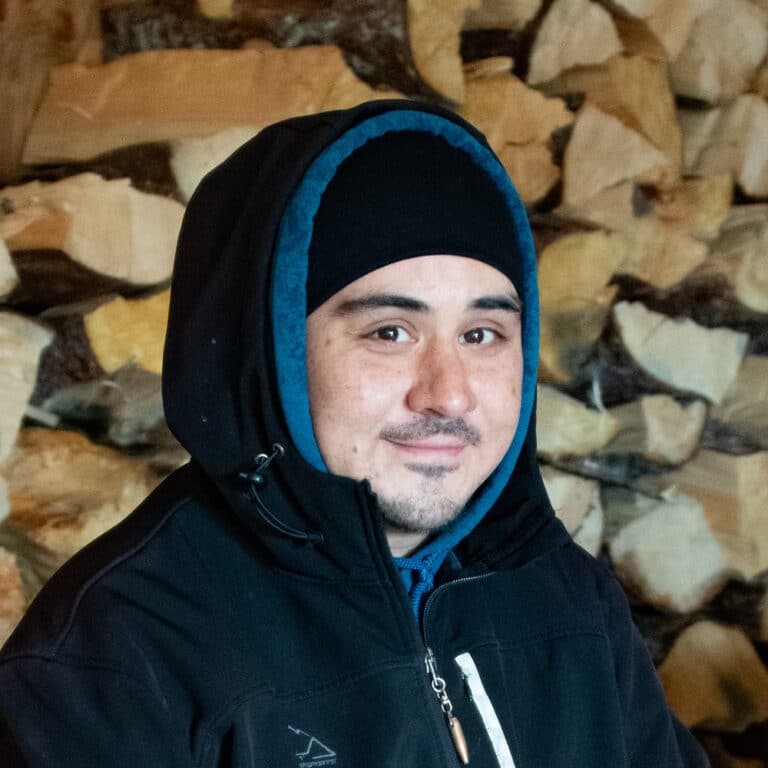 Man wearing a hat and hood in front of a stack of fire wood